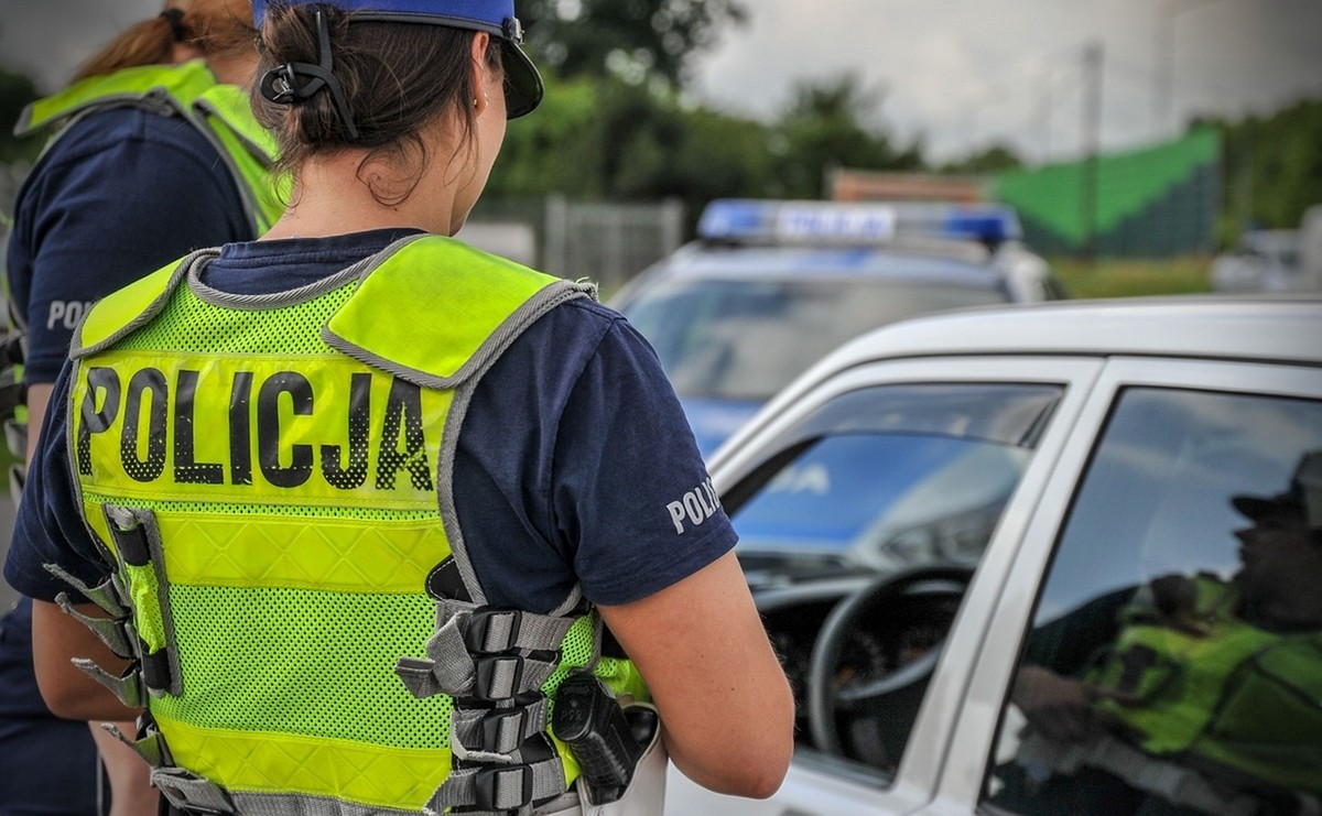 Policjanci w kamizelkach odblaskowych z napisem rozmawiają z kierowcą zatrzymanego samochodu. W bagażniku samochodu ujawniono sześciu migrantów.