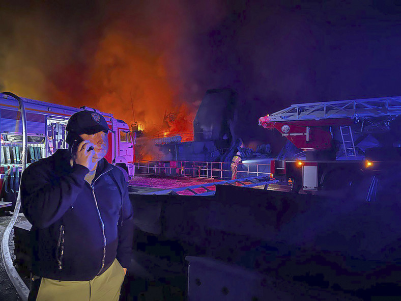 Mężczyzna rozmawiający przez telefon, w tle dym i płomienie unoszące się z płonącej stoczni w Sewastopolu na Krymie, 13 września 2023 r.