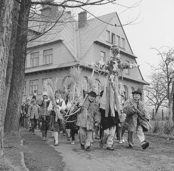 Procesja dzieci w strojach góralskich niosących kukłę Marzanny. Jedno z dzieci niesie sanki, drugie narty. W tle dom wielorodzinny (NAC, domena publiczna, sygnatura 3/53/0/6/341).