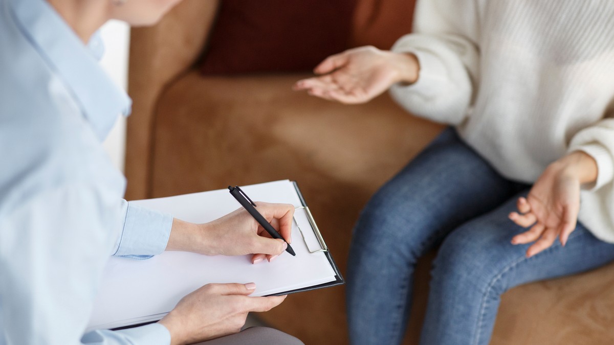 Osoba prowadząca notatki na kartce rozmawia z drugą osobą siedzącą naprzeciwko, która gestykuluje rękami — scena przypomina spotkanie terapeutyczne lub rozmowę o pracę.