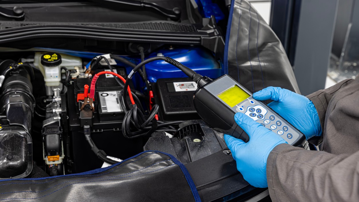 Vodič s otvorenou kapotou automobilu, ktorý sa snaží diagnostikovať problém s motorom pomocou základného náradia a multimetra na kontrolu napätia batérie. 
