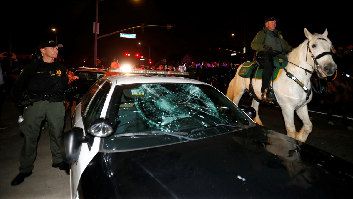 Police on horseback and in riot gear pass by a damaged police car as they break up a demonstration outside Republican U.S. presidential candidate Donald Trump's campaign rally in Costa Mesa