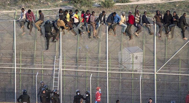 African migrants sit atop a border fence as Spanish police in riot gear try to turn