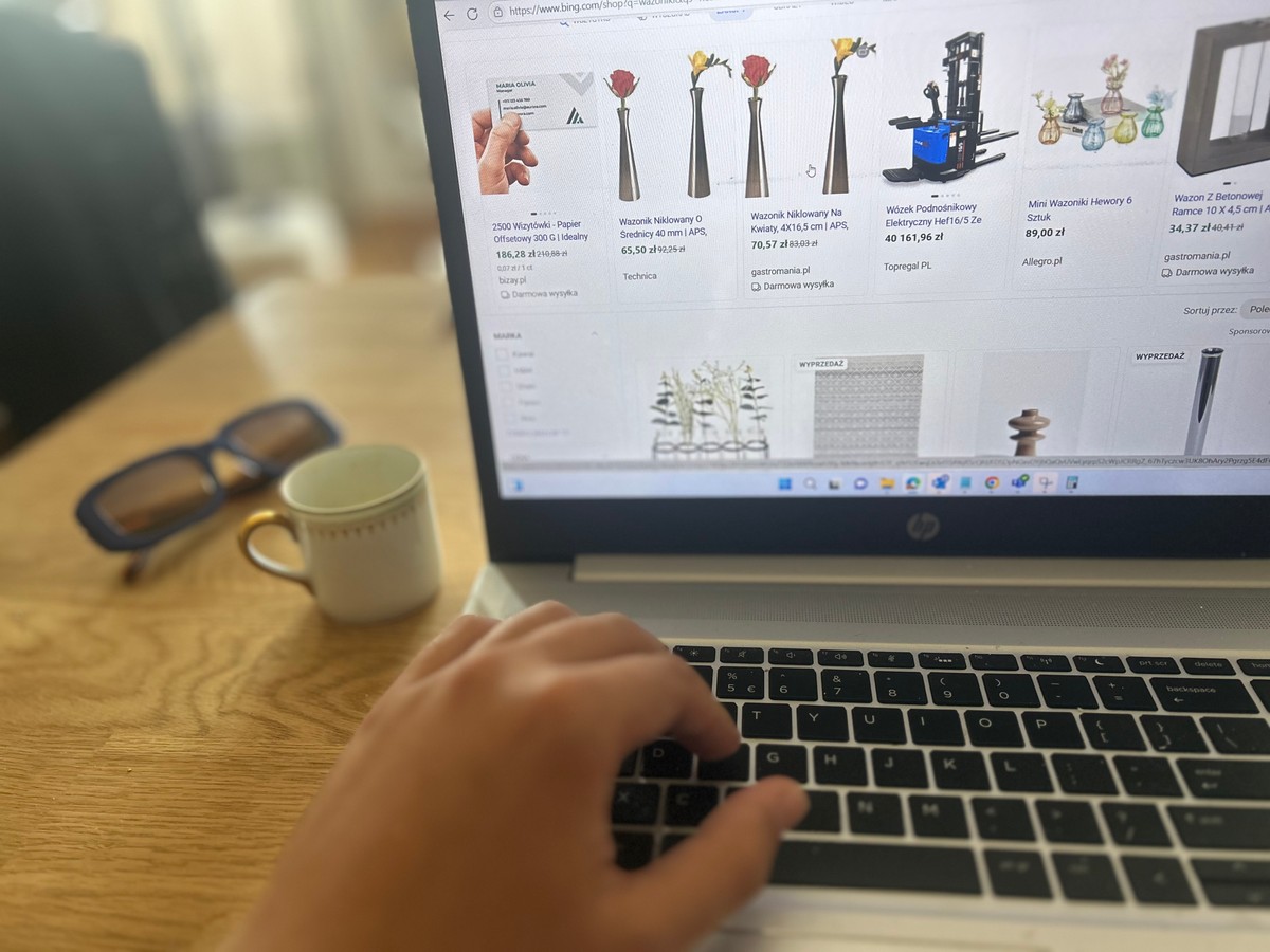 Osoba korzysta z laptopa HP, przeglądając ofertę zakupów online z wazonami na stronie internetowej. Na drewnianym stole obok komputera leżą okulary przeciwsłoneczne i mała filiżanka.
