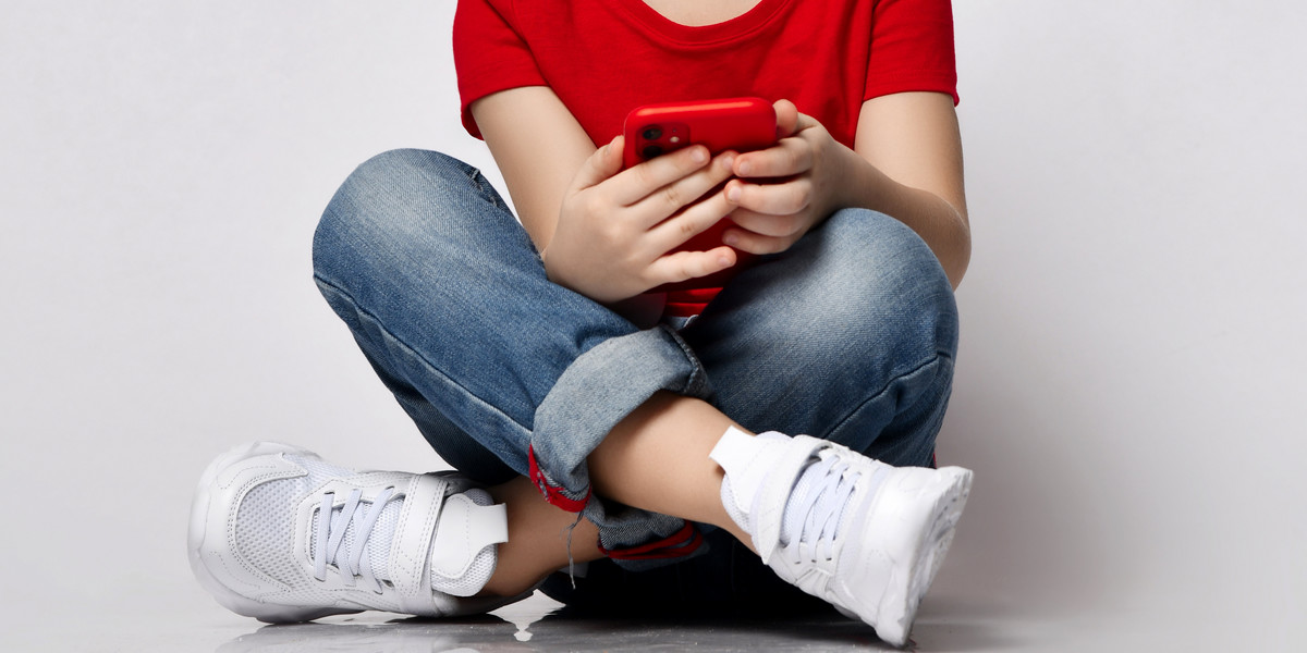 Stylish kid boy child in red t-shirt, jeans, sneakers and sunglasses sitting on floor with legs cros