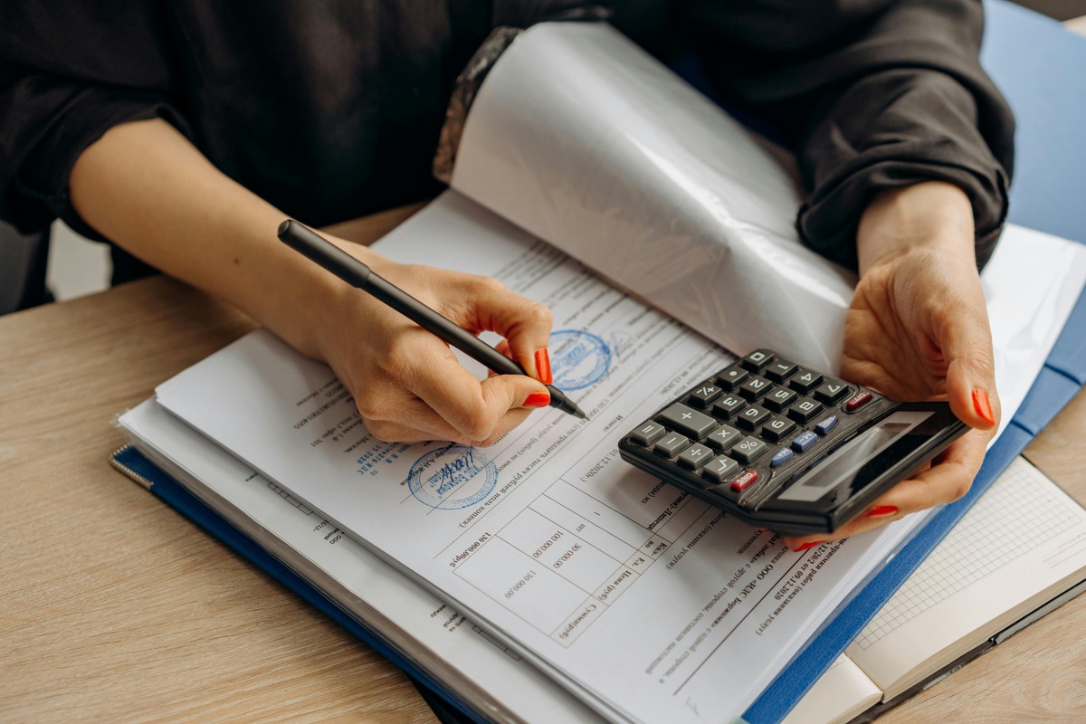 Osoba trzymająca kalkulator i jednocześnie podpisująca dokumenty finansowe na biurku, obok segregatora i stosu papierów.