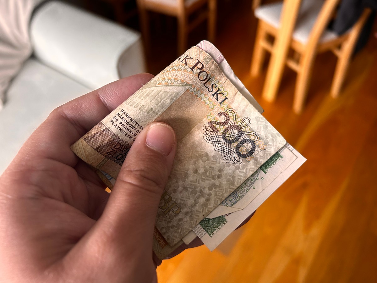 Zbliżenie na dłoń trzymającą banknoty polskie, w tym widoczny banknot o nominale 200 zł. W tle rozmyte wnętrze mieszkania z drewnianymi krzesłami i stołem.
