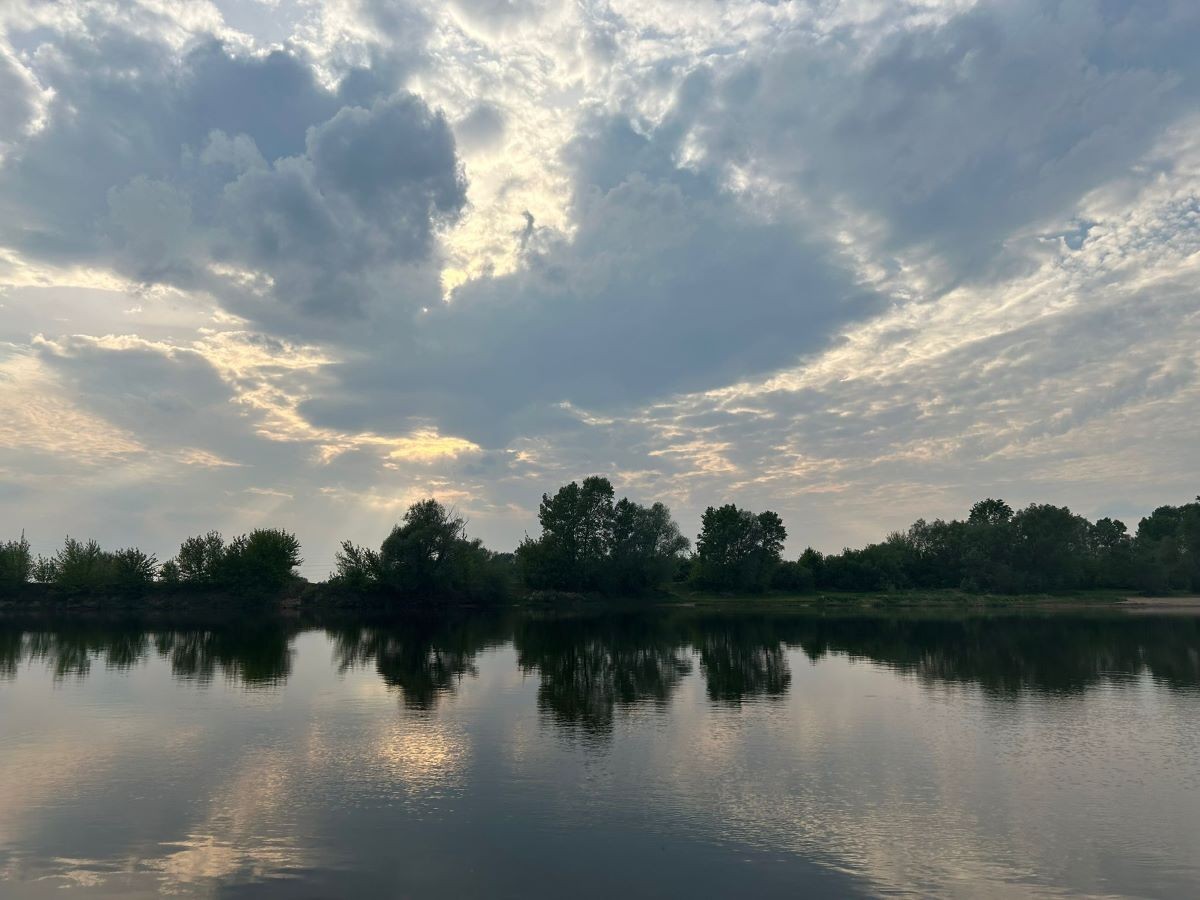 Zachmurzone niebo o świcie odbijające się w spokojnej tafli jeziora, otoczonego zielonymi drzewami.