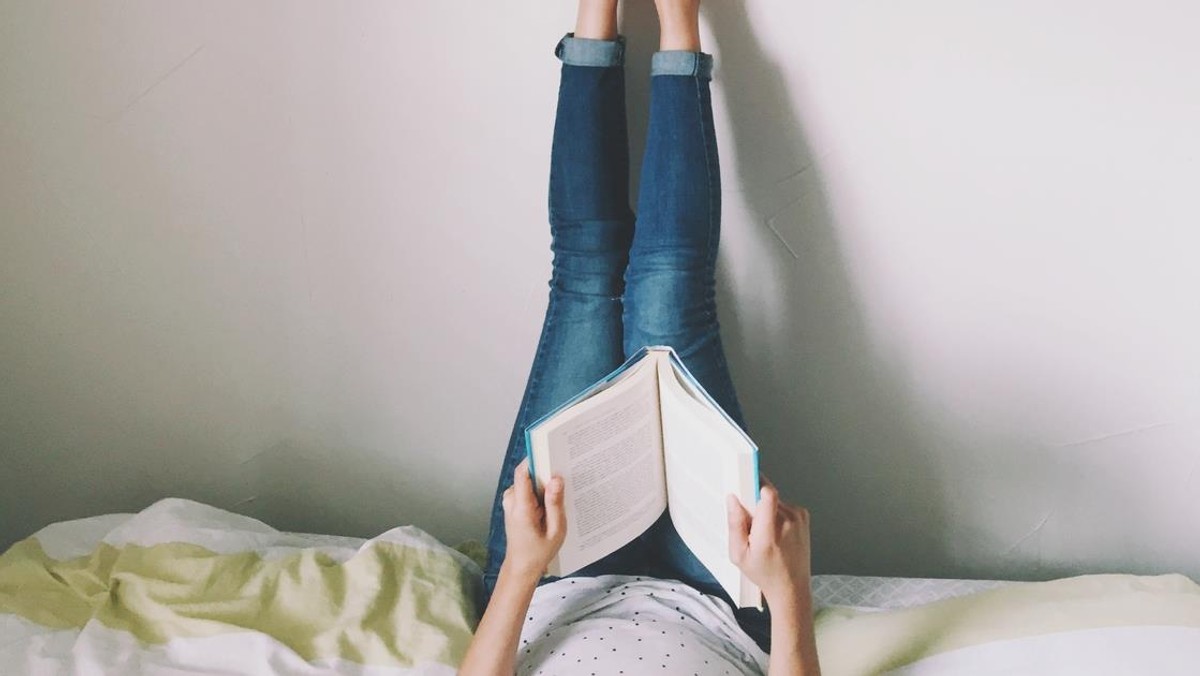 Woman Lying On Bed Reading Book With Legs Raised