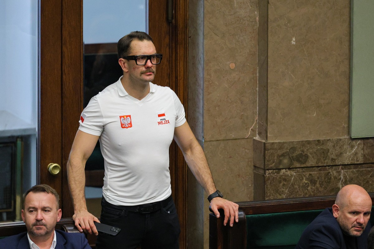 Poseł Mejza ubrany w białą koszulkę polo z napisem „Poseł Mejza” oraz godłem Polski stoi w ławach sejmowych, opierając się o poręcz. Wokół niego siedzą inni parlamentarzyści, a tło stanowi marmurowa ściana sali sejmowej.