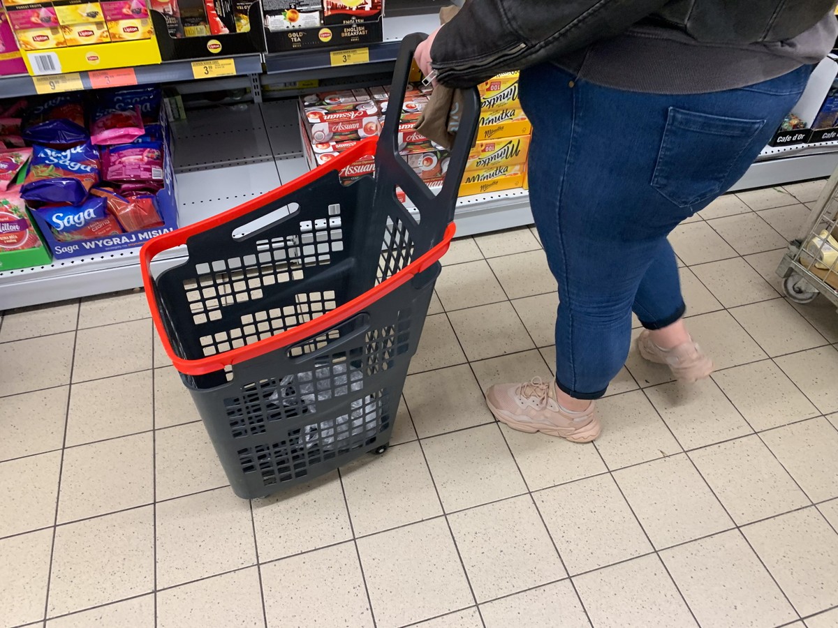Osoba w dżinsach i sportowych butach prowadzi koszyk na kółkach w alejce sklepu spożywczego, obok półek z herbatą i innymi produktami.