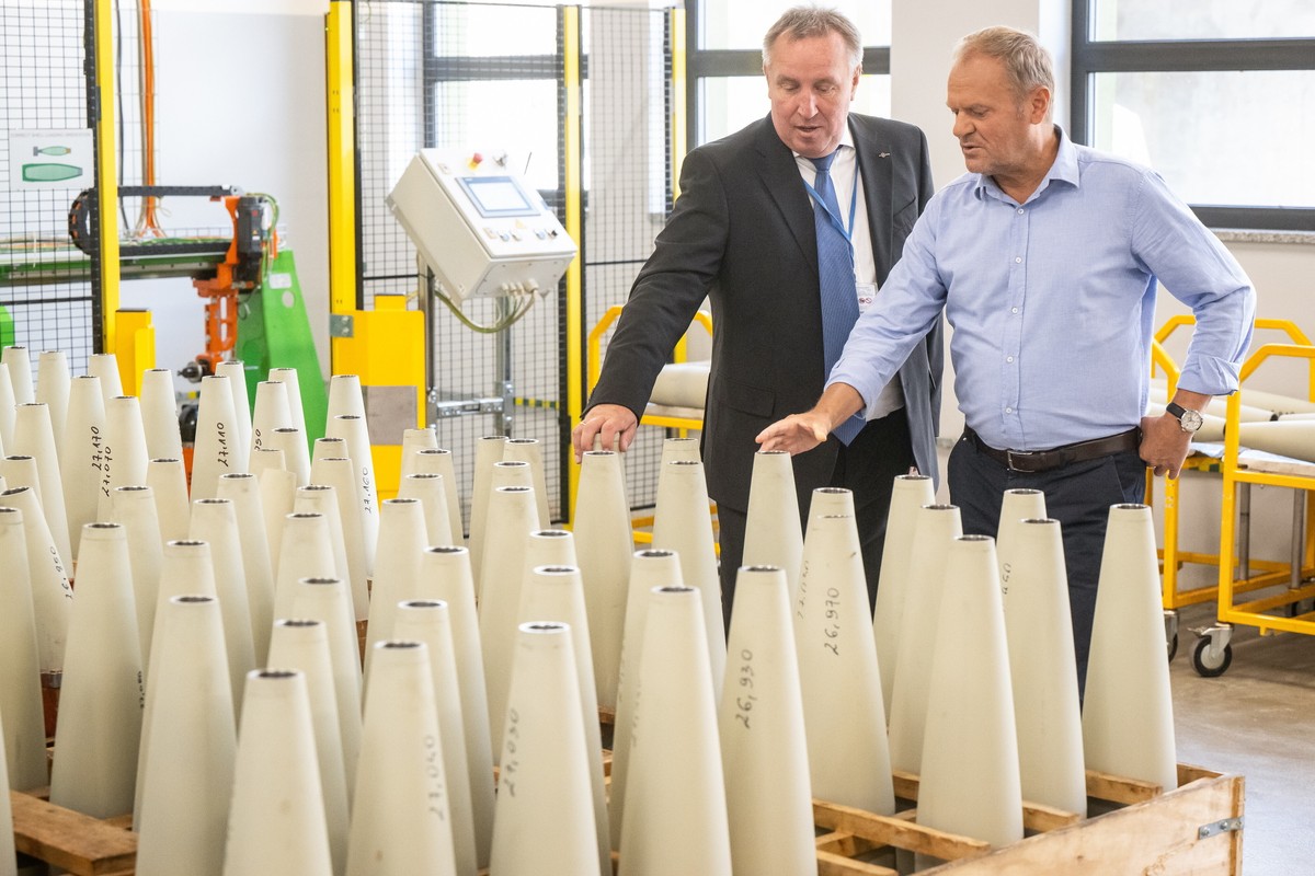 Na zdjęciu Donald Tusk i Arkadiusz Miszuk w garniturze i koszuli stoją w hali produkcyjnej, oglądając rzędy jasnych korpusów pocisków ustawionych na paletach; w tle widoczne są maszyny i elementy linii technologicznej.