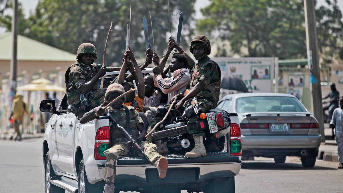 Soldiers and people carrying machetes ride on the back of a vehicle along a street in Gombe