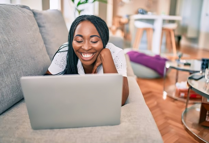Young woman smiling while working using laptop laying on the sofa at home