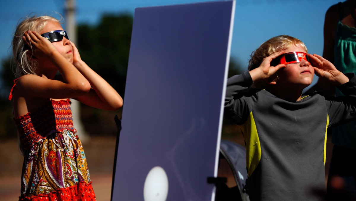 A reflected image of the sun is seen on a white board as kids look up to view the beginning a partia