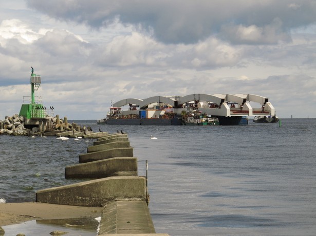 Dla portu w Gdańsku jednotorowy most nad Martwą Wisłą jest dziś wąskim gardłem. Z powodu złego stanu technicznego obowiązuje tu ograniczenie prędkości dla pociągów towarowych do 10 km/h. Nowa szersza przeprawa ma udrożnić obsługę kolejową największego polskiego portu. W piątek na barkach dopłynęły ze stoczni Nauta w Gdyni stalowe łukowe przęsła nowej przeprawy. Transport zacumował w rejonie Trasy Sucharskiego. Dzisiaj trwa ustawianie potężnego dźwigu, którego zadaniem będzie umieszczenie przęseł na przyczółkach. Jak zapowiada spółka PKP Polskie Linie Kolejowe, ten etap powinien się zacząć w połowie lipca.