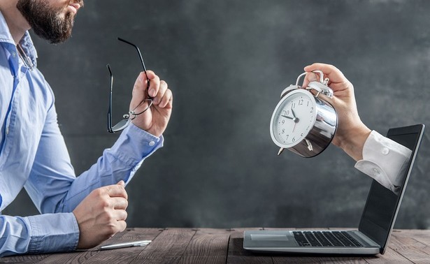 Businessman is sitting at the desk and is looking at the hand with clock coming out of the laptop. Metaphor of spending time at work