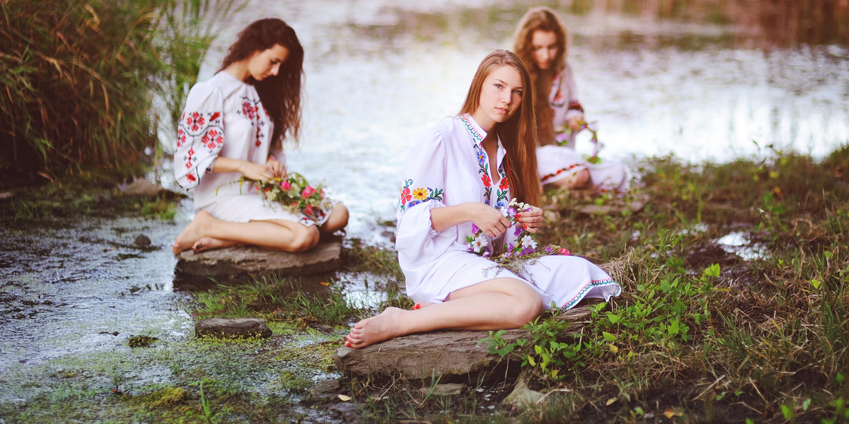 three young beautiful girls in white shirts with floral ornaments with wreaths in their hands sitting on the background of the river.