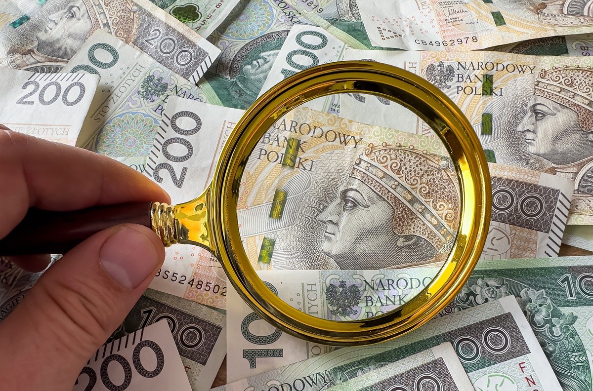 Dłoń trzymająca złotą lupę nad banknotami 100 i 200 zł