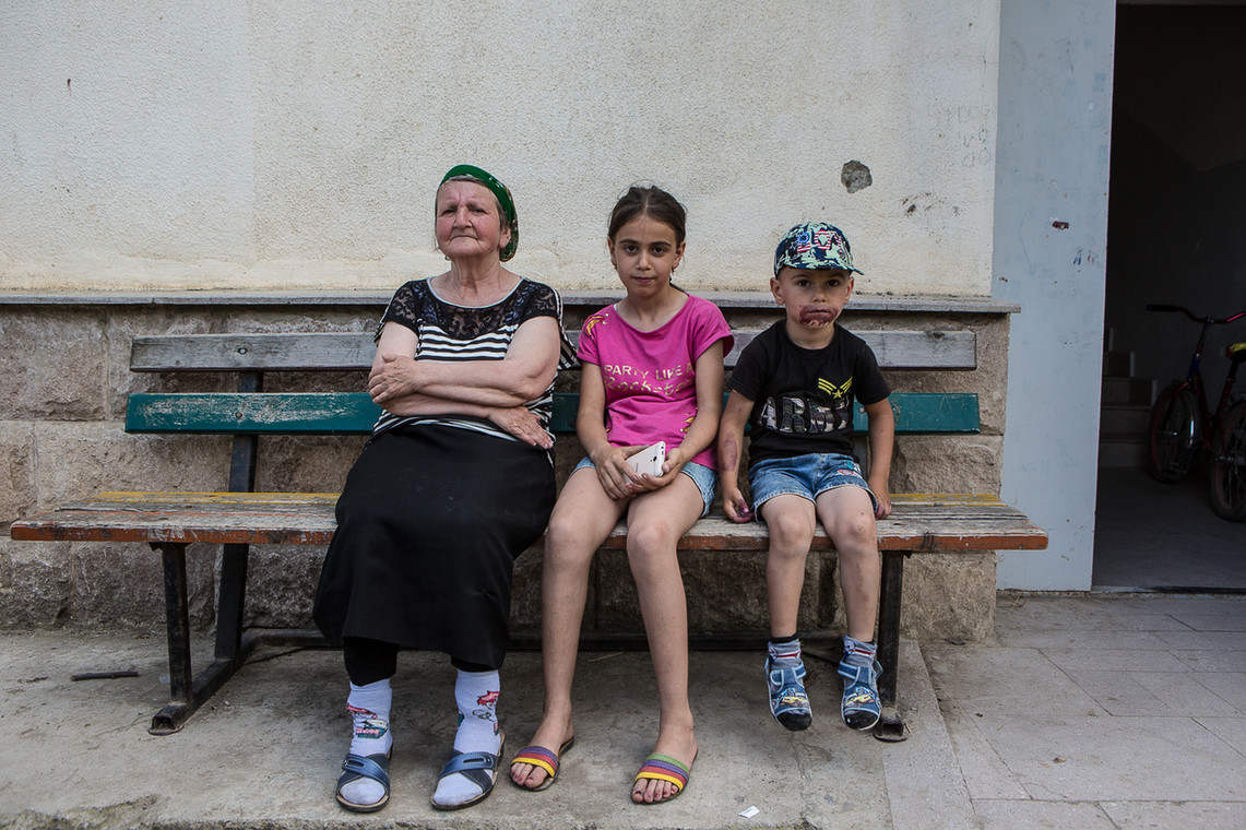 Babcia siedząca z wnuczętami przed jednym z bloków. Nad głową chłopca ślad po kuli