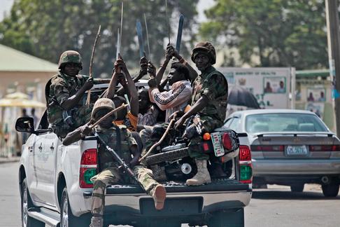 Soldiers and people carrying machetes ride on the back of a vehicle along a street in Gombe