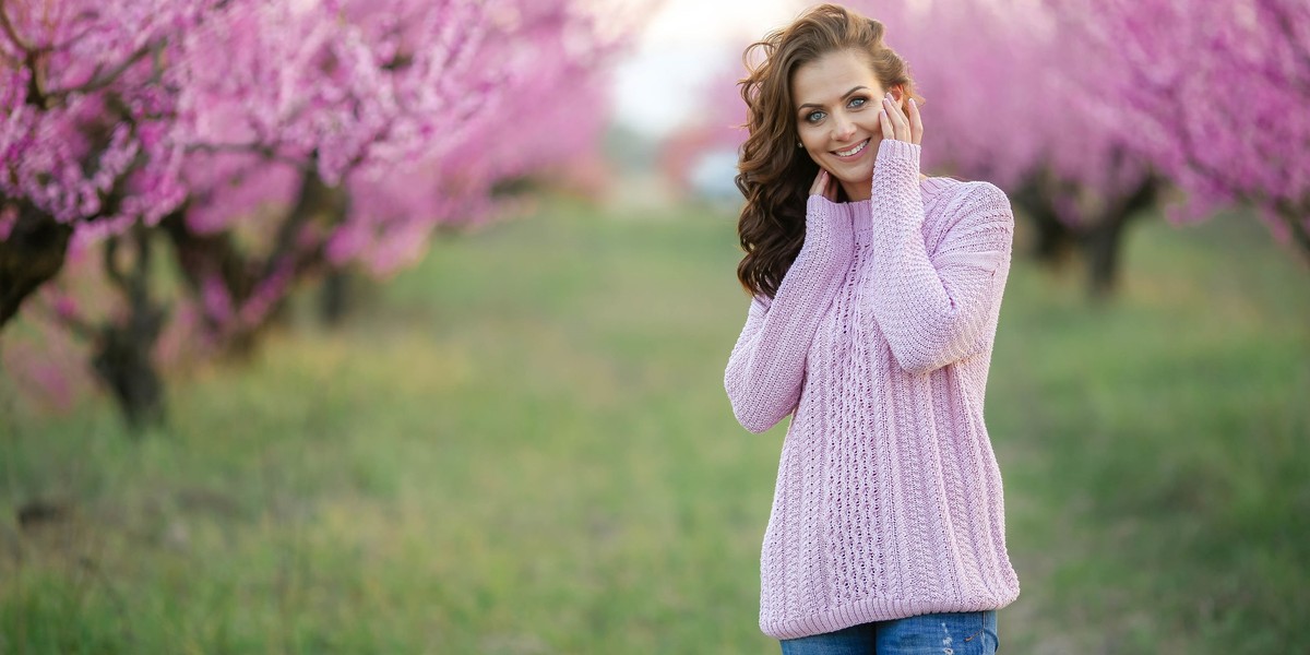 a stylish adult woman in a glamorous sweater near the branches of a tree covered with pink blossoms