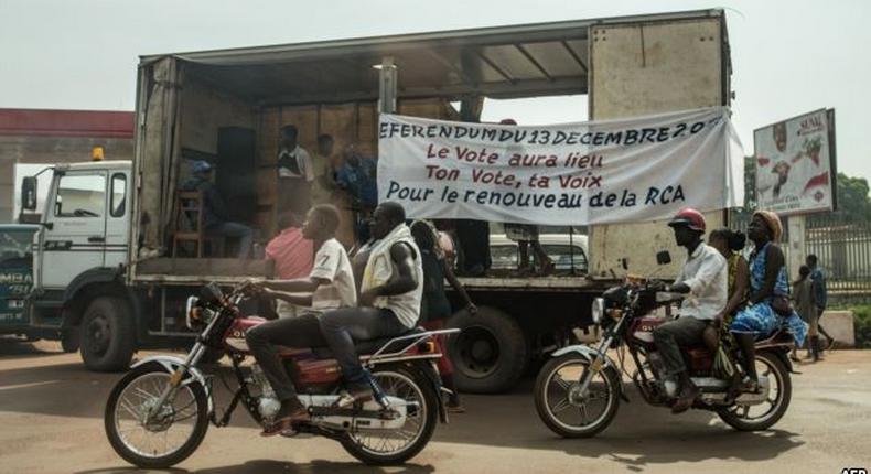 Motorbikes past an electoral caravan with a banner that reads, The vote will take place, your vote, your voice, in Bangui, Central African Republic.