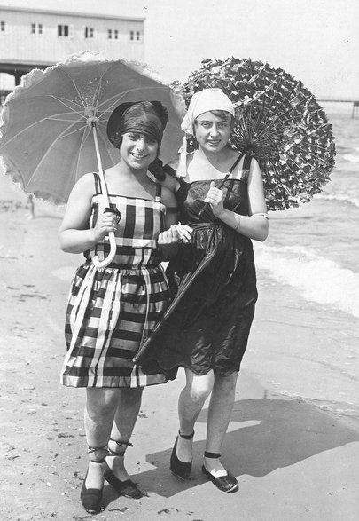 Kobiety w strojach kąpielowych i nakryciach głowy z parasolkami przeciwsłonecznymi podczas spaceru po plaży nad Morzem Bałtyckim