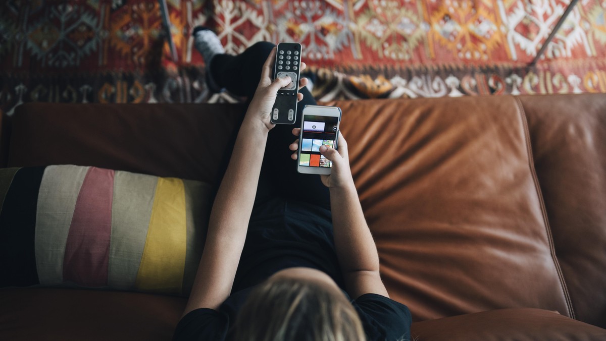 High angle view of teenage girl using phone app and remote control while sitting on sofa watching TV