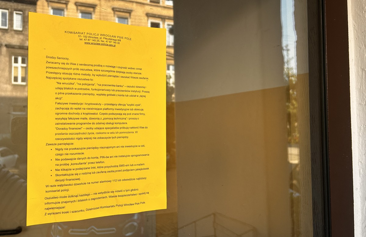 Żółta kartka z komunikatem Komisariatu Policji Wrocław Psie Pole przyklejona na szybie budynku. Treść skierowana do seniorów ostrzega przed oszustwami metodą „na wnuczka”, „na policjanta”, fałszywymi inwestycjami i podszywaniem się pod doradców finansowych. Zawiera wskazówki, jak unikać wyłudzeń, np. by nie przekazywać pieniędzy obcym, nie podawać danych do konta ani nie klikać w podejrzane linki.