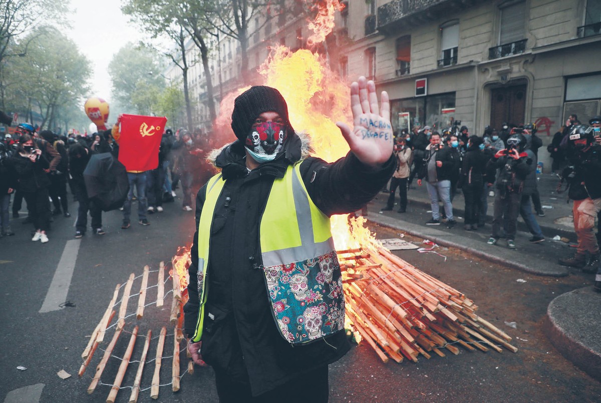 Demonstrant w żółtej kamizelce z zasłoniętą twarzą podnosi rękę w geście stop, za nim płoną barykady, a w tle widać tłum protestujących na ulicy.