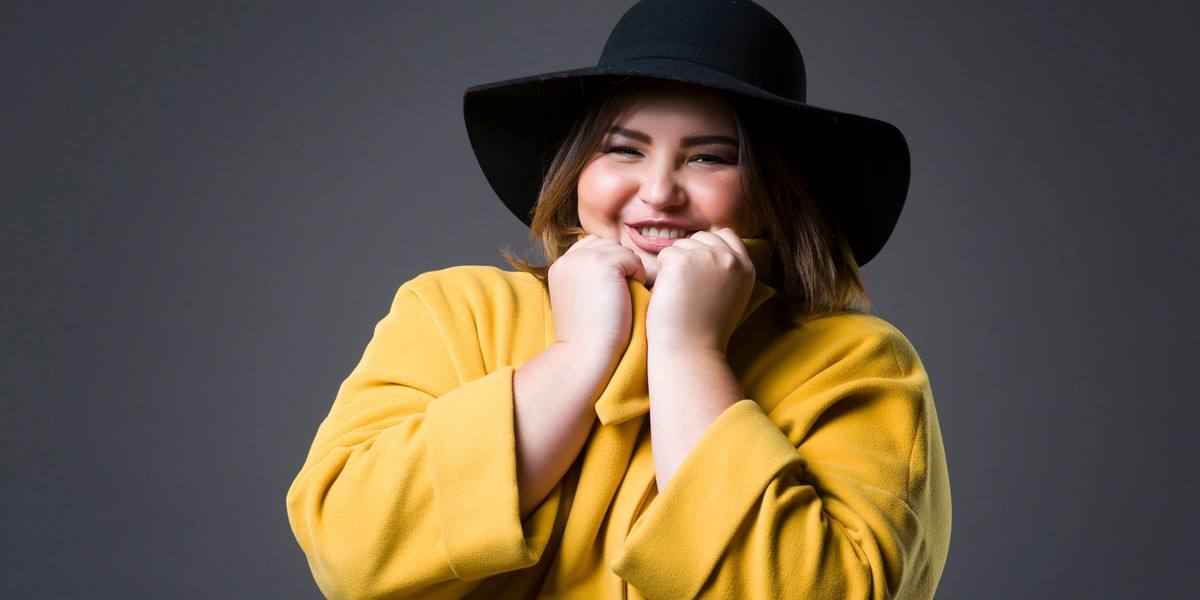 Plus size fashion model in yellow coat and black hat, fat woman on gray background, overweight femal