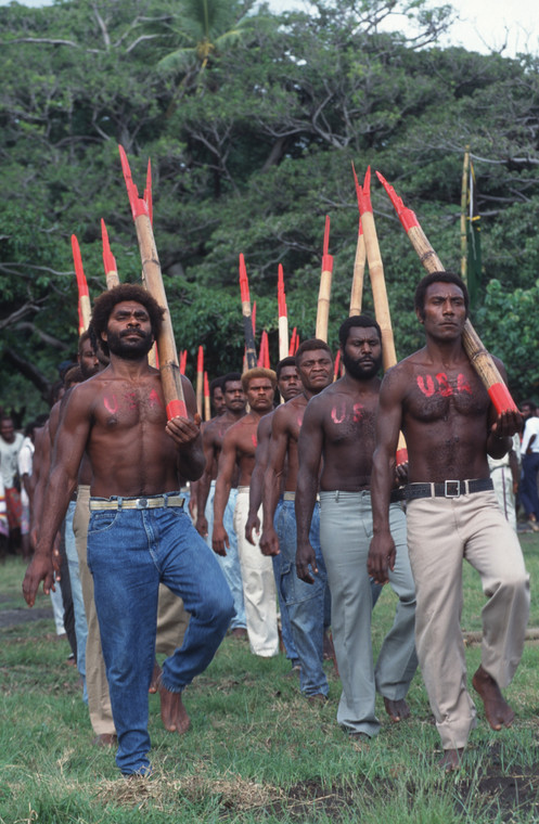 Mieszkańcy Vanuatu podczas parady na cześć Johna Fruma. Mężczyźni trzymają w rękach drewniane kije, które imitują karabiny, a na swoich torsach mają napis "USA"