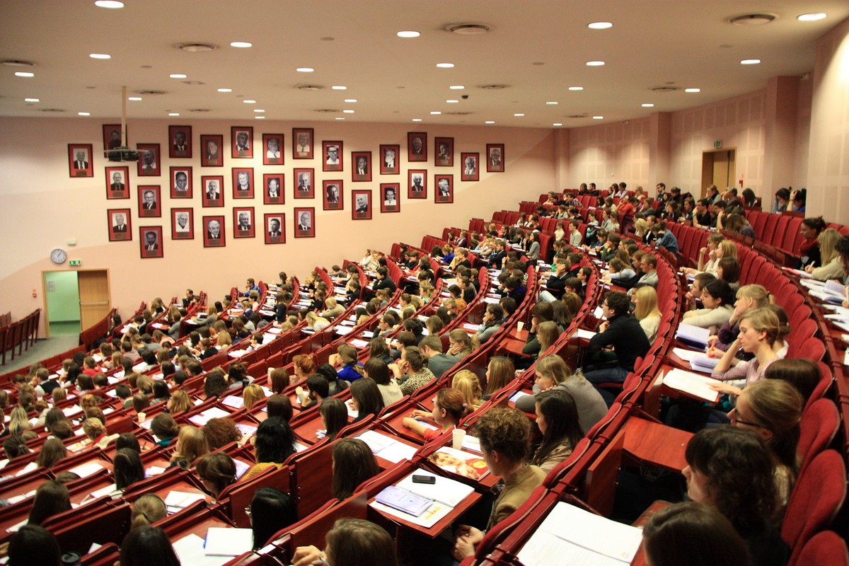 Duża aula uniwersytecka z czerwonymi siedzeniami, w której trwa wykład. Studenci siedzą rzędami, notując w zeszytach. Na tylnej ścianie widoczne są liczne portrety, prawdopodobnie przedstawiające byłych rektorów lub zasłużonych wykładowców.