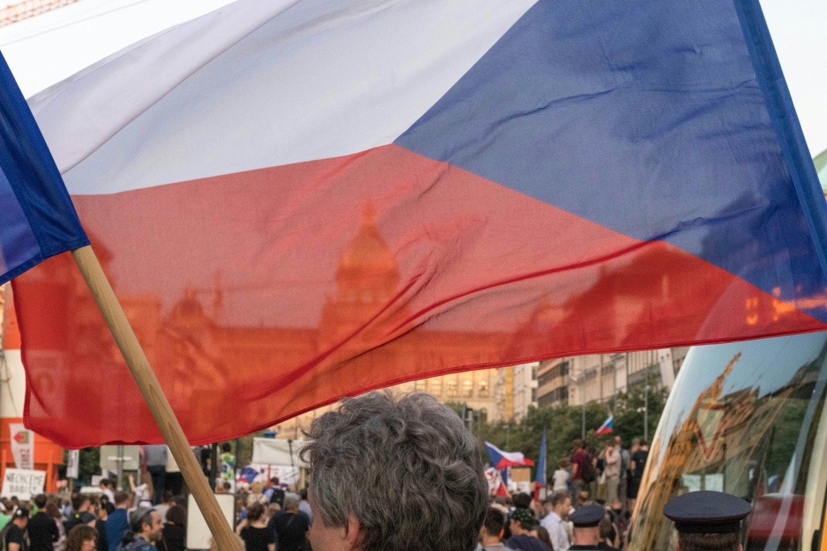 Duża flaga Czech trzymana na kiju powiewa nad tłumem ludzi podczas zgromadzenia w przestrzeni miejskiej, w tle widoczny zarys zabytkowego budynku i inne czeskie flagi.