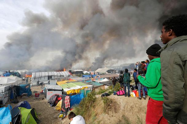 Migrants look at burning makeshift shelters and tents in the Jungle on the third day of their evac