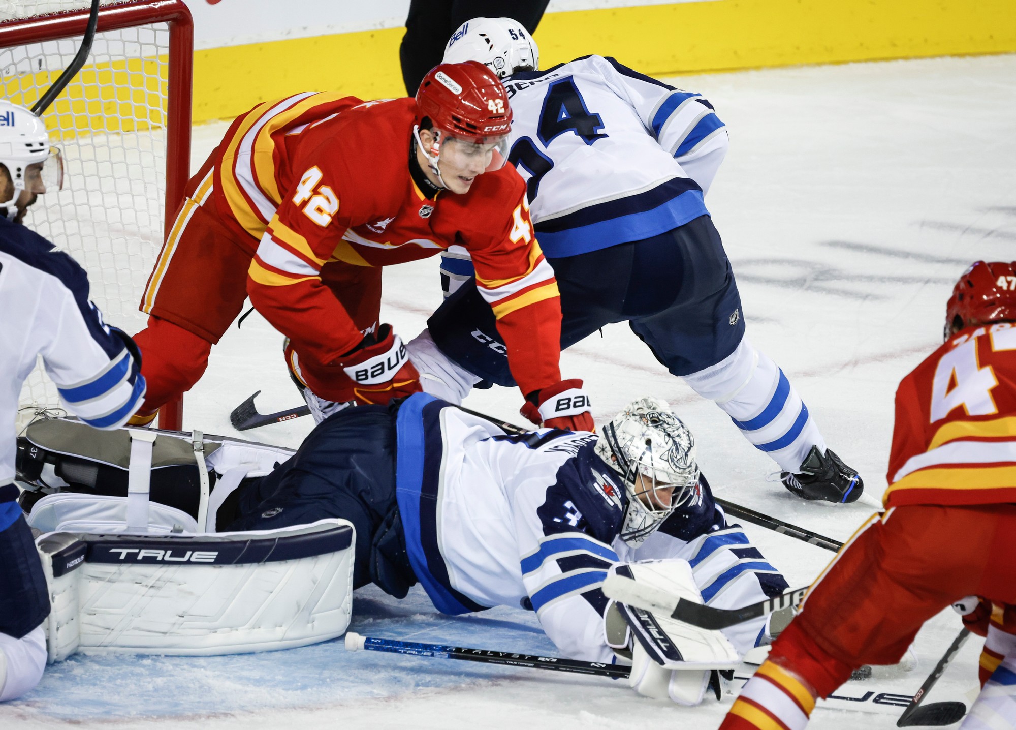 Slovenský hokejista v drese Calgary Flames Samuel Honzek (42), brankár Winnipegu Jets Connor Hellebuyck (uprostred) a jeho spoluhráč Dylan Samberg (druhý sprava).