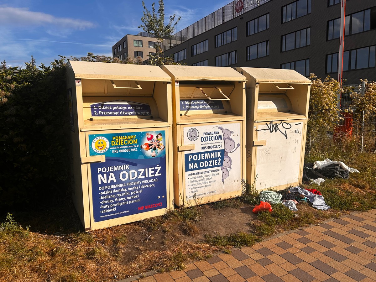 Dwa żółte pojemniki na odzież stojące przy chodniku, oznaczone napisami „Pojemnik na odzież – Pomagamy dzieciom”. Na ziemi obok leży porozrzucana odzież, a jeden z pojemników jest pomazany graffiti. W tle widać krzewy oraz nowoczesny budynek biurowy.