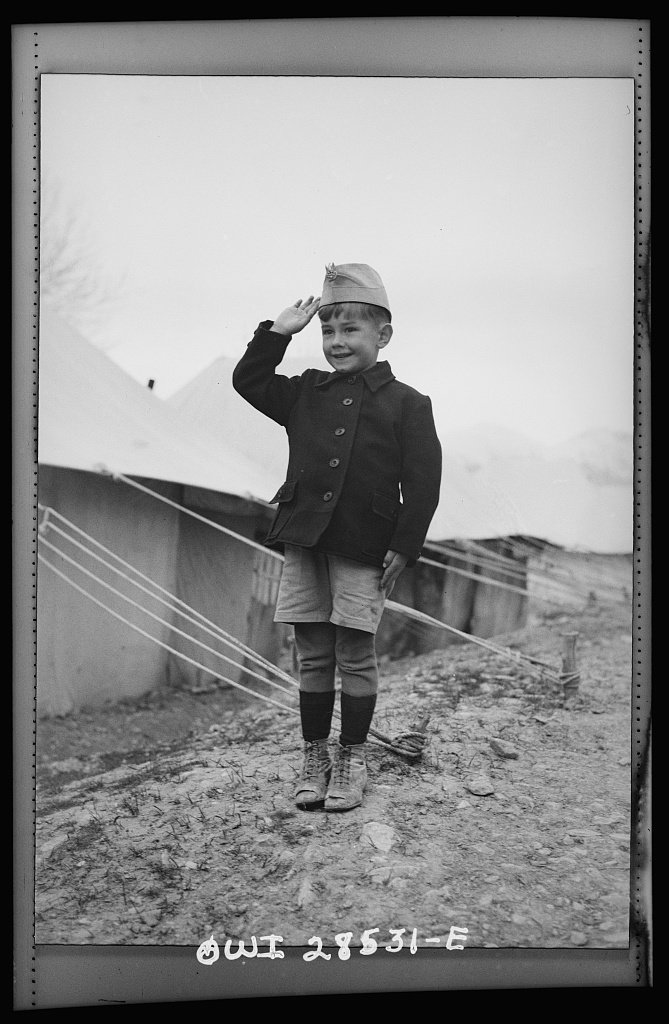 Chłopiec ubrany w marynarkę i wojskową czapkę, w tle namioty obozowe (domena publiczna)