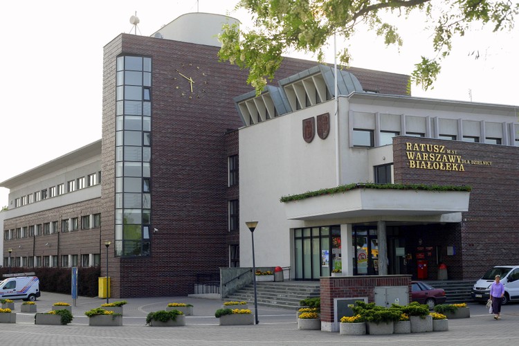 Drugie miejsce zajęły ex aequo Białołęka i Wesoła z 59 proc. odsetkiem wskazań. W przypadku Białołęki jest to porównywalny wynik do ubiegłorocznego rankingu. Jak mantra w komentarzach powtarzana jest niekorzystna lokalizacja dzielnicy (nawet pomimo nowej linii tramwajowej, coraz lepszych połączeń z metrem i innymi dzielnicami) oraz brak dostępnej (zwłaszcza na tzw. zielonej Białołęce) infrastruktury. Wraz z kolejnymi inwestycjami w tej dzielnicy można oczekiwać, że stereotypowy wizerunek będzie się zmieniał na lepsze. Już wkrótce Białołęka zyska np. galerię handlową, co pozbawi przeciwników zamieszkiwania w tej dzielnicy pewnej liczby negatywnych argumentów. Na razie, wbrew wynikom ankiet, jest to miejsce, w którym intensywnie rozwija się budownictwo deweloperskie, to właśnie tu są największe szanse na dopłatę w ramach Mieszkania dla Młodych, a dzielnica zamyka pierwszą piątkę najczęściej wybieranych lokalizacji w Warszawie na rynku wtórnym. Nie może to dziwić, jeżeli spojrzymy na ceny w transakcjach (średnio 6385 zł za m kw.), podczas gdy średnia warszawska jest o 1000 zł wyższa. <br><br>
Na zdj. Ratusz dzielnicy Białołęka - fot. Cezary p - licencja CC BY-SA 3.0