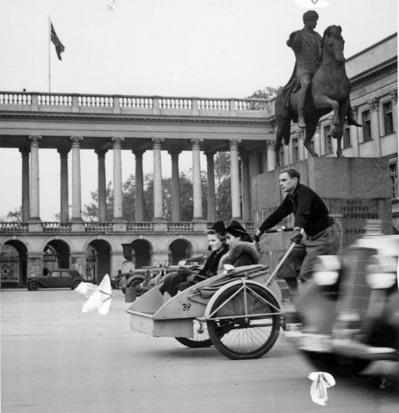 Przedwojenna riksza z dwoma kobietami przejeżdża przez pl. Saski. Widoczny Pałac Saski i pomnik ks. Józefa Poniatowskiego