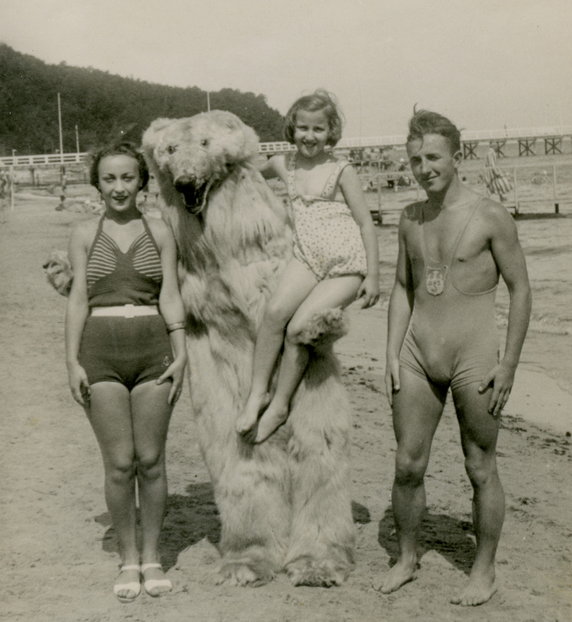 Zdjęcie z lat 1935-39 na plaży w Orłowie. Po prawej stronie  młody mężczyzna w kostiumie kąpielowym z naszytą tarczą Akademickiego Związku Sportowego. W tle drewniane molo i zalesiony cypel Kępy Redłowskiej.  