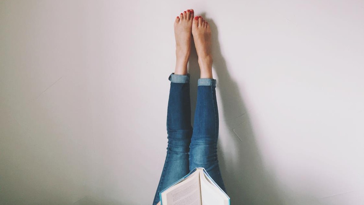 Woman Lying On Bed Reading Book With Legs Raised