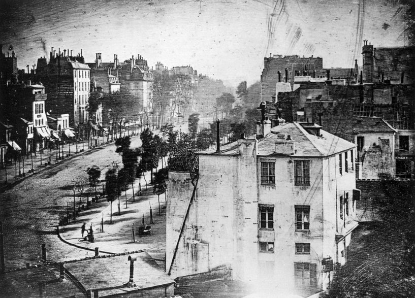 Louis Daguerre, Boulevard du Temple w Paryżu, pierwsza fotografia z widocznym żywym człowiekiem (w lewym dolnym rogu), który zatrzymał się na wystarczająco długo, by zostać zarejestrowanym przez fotografa (1838, domena publiczna)