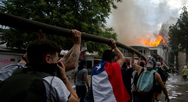 Demonstrators hold up a hose as flames engulf the Pedro de Valdivia University following a protest against the government of Chilean President Sebastian Pinera in Santiago, on November 8, 2019.Unrest began in Chile last October 18 with protests against a rise in transport tickets and other austerity measures that descended into vandalism, looting, and clashes between demonstrators and police.