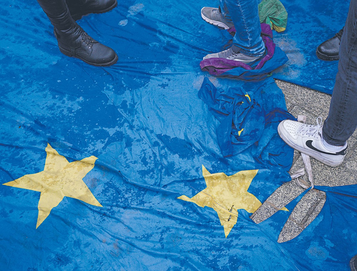 Fragment dużej flagi Unii Europejskiej leżącej na ziemi, po której stoją i chodzą ludzie w butach, częściowo zasłaniając żółte gwiazdy na niebieskim tle.