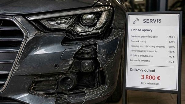 Detail rozbitého predného svetlometu moderného auta po parkovacom náraze – viditeľný poškodený nárazník s radarovým senzorom a kamerou, vedľa ceduľka servisu s odhadom opravy 3 800 eur.