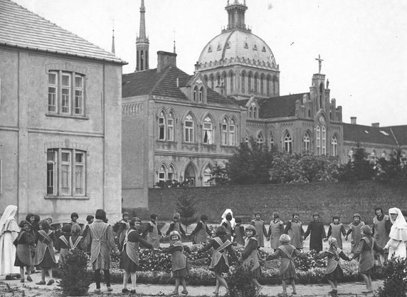 Rok 1928. Dzieci z przyklasztornej ochronki i szkoły w Płocku bawią się w ogrodzie przyszkolnym. W tle Świątynia Miłosierdzia i Miłości. Zdjęcie z Narodowego Archiwum Cyfrowego, zespół: Koncern Ilustrowany Kurier Codzienny - Archiwum Ilustracji. Sygnatura: 3/1/0/15/1058