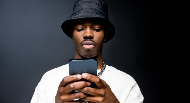 Portrait of handsome young man wearing white sweatshirt and black bucket hat, using mobile phone. Studio shot on black background. 
