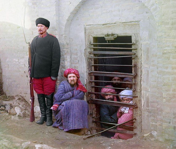 Zindan (tradycyjne więzienie w Azji Centralnej). Więźniowie patrzą przez kraty, pilnuje ich strażnik ubrany w rosyjski mundur. Prawdopodobnie zdjęcie zrobione w Bucharze w Użebkistanie (między 1905 i 1915, domena publiczna).